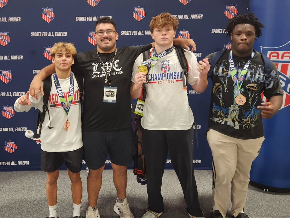 Coach Ray Lugo, in a black Lugo Wrestling Club shirt and a coach’s pass, with three wrestlers wearing medals in front of an AAU backdrop.