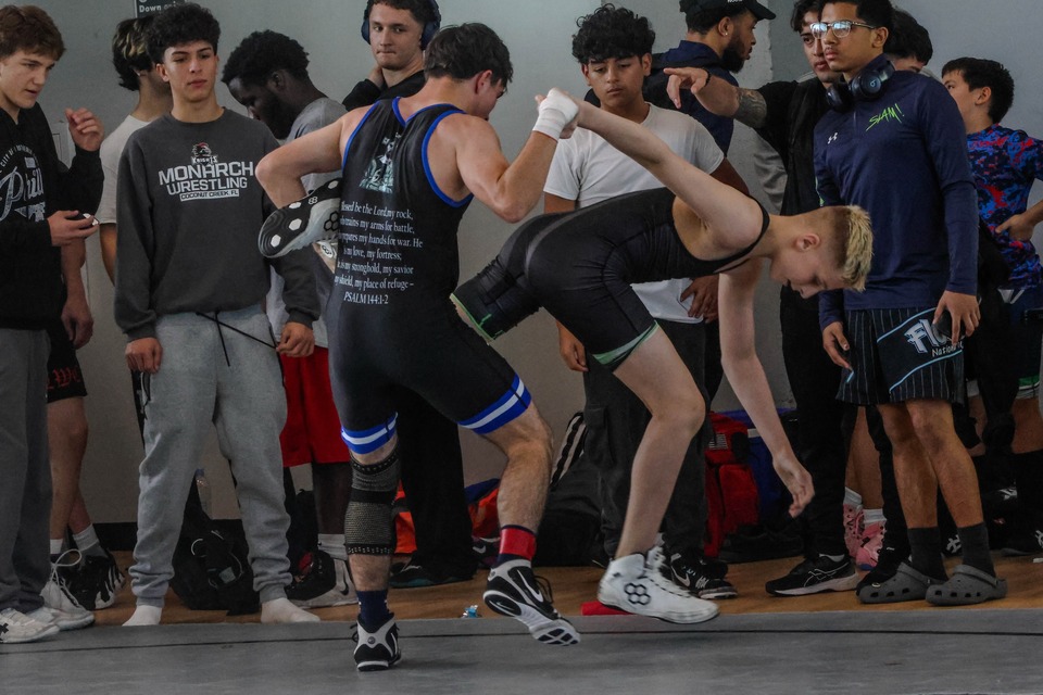 An LWC wrestler in a black singlet lifts an opponent’s leg at a tournament while other wrestlers watch from the edge of the mat.